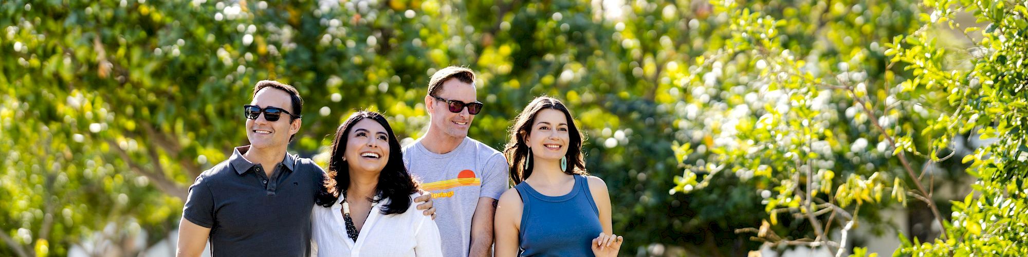 Four friends pose outdoors in a sunny orchard, smiling and carrying shopping bags with mountains in the background.