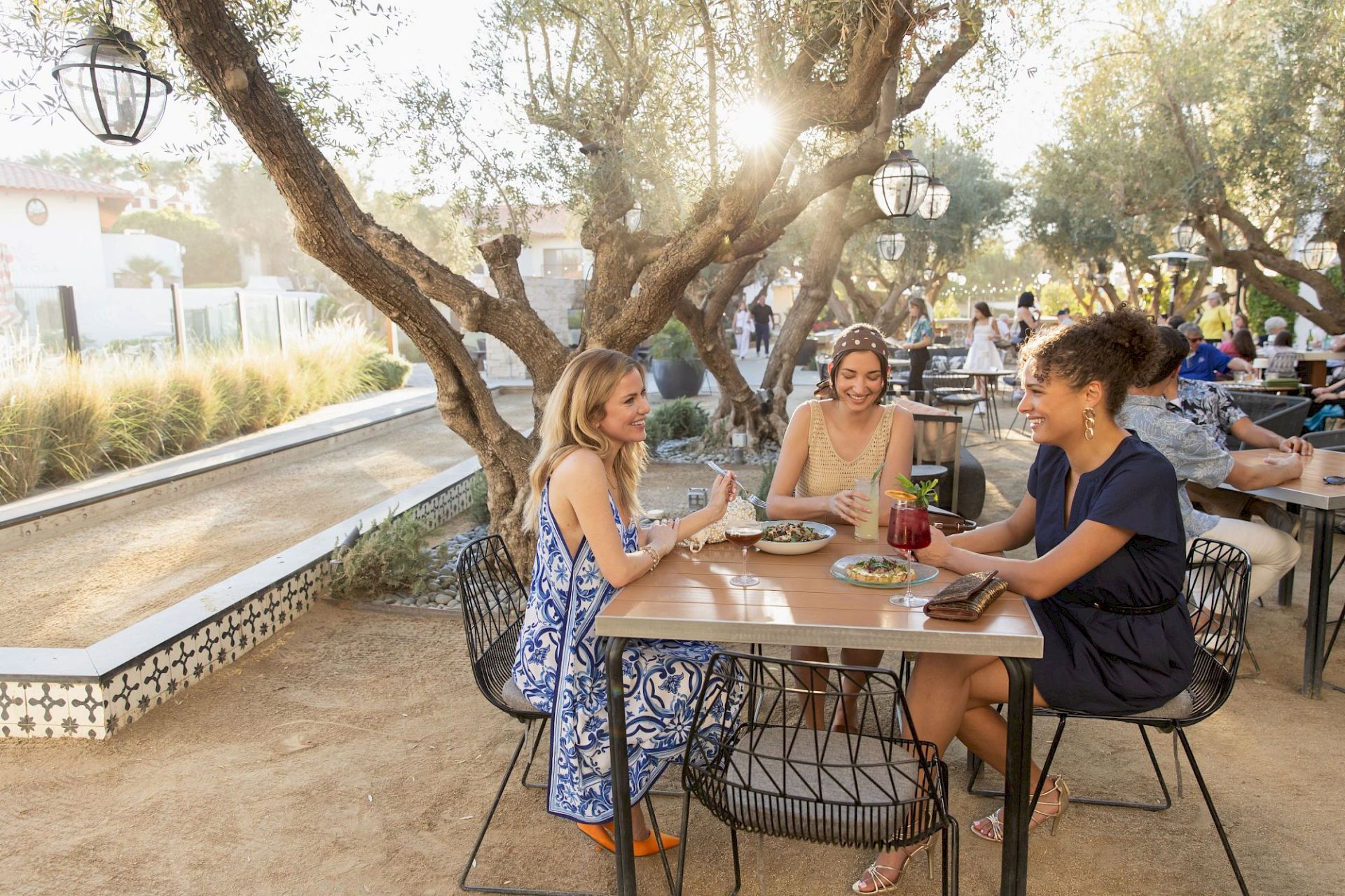 Friends dine outdoors at a shaded patio, laughing over drinks and plates beneath lantern-lit trees.