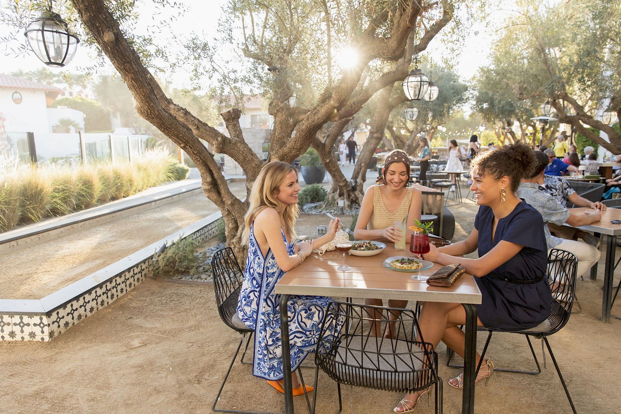 Friends dine outdoors at a shaded patio, laughing over drinks and plates beneath lantern-lit trees.