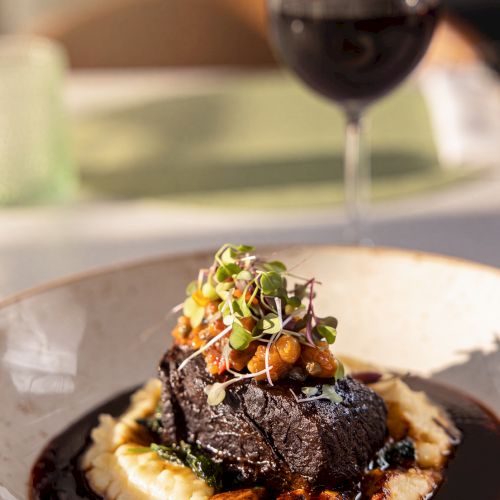 A fancy plated entr&eacute;e with steak, creamy potatoes, microgreens, and a glass of red wine in the background.