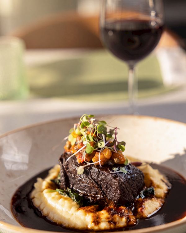 A fancy plated entr&eacute;e with steak, creamy potatoes, microgreens, and a glass of red wine in the background.