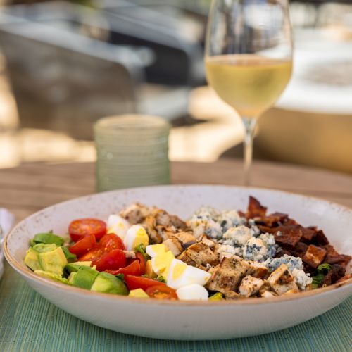 A colorful salad with grilled meats and avocado sits on a table, with a glass of white wine in the background.