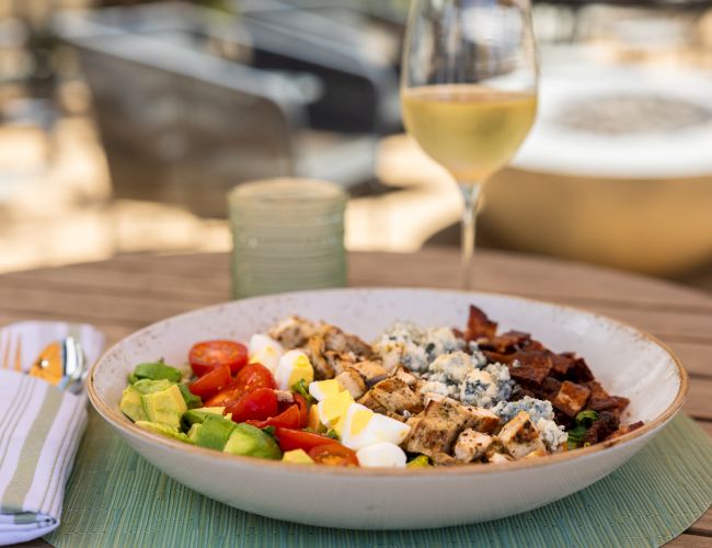 A colorful salad with grilled meats and avocado sits on a table, with a glass of white wine in the background.