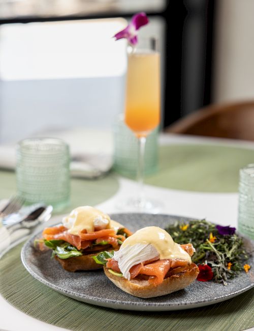 A plated open-faced sandwich with smoked salmon and greens sits beside an orange drink in a glass.