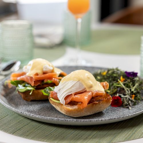 A plate of open-faced toast topped with smoked salmon, fresh greens, and soft cheese, with bowls and glasses blurred in the background.