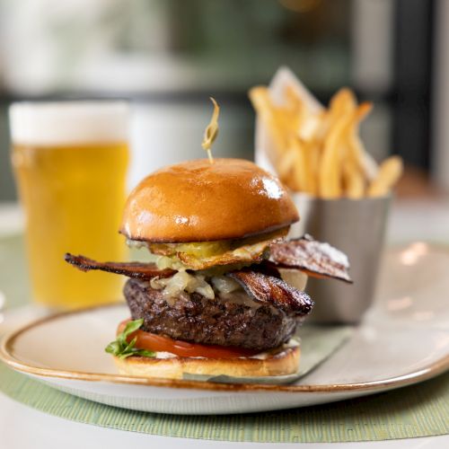 A juicy burger with cheese and bacon sits on a plate, with fries and a glass of orange drink in the background.