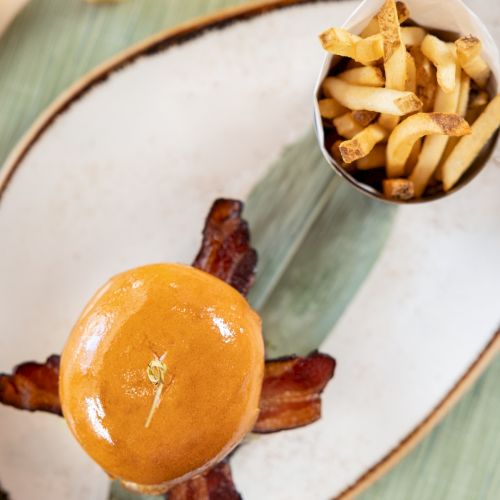 Close-up of fries in a cup beside a glossy burger bun on a plate with bacon and a small drink.