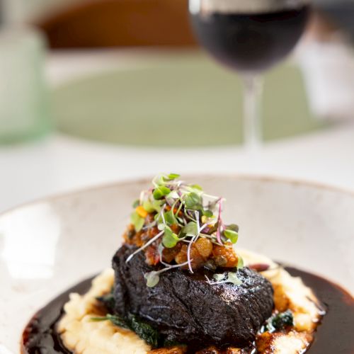 A plated dark meat steak with creamy mash and colorful microgreens, with a glass of red wine softly blurred in the background.