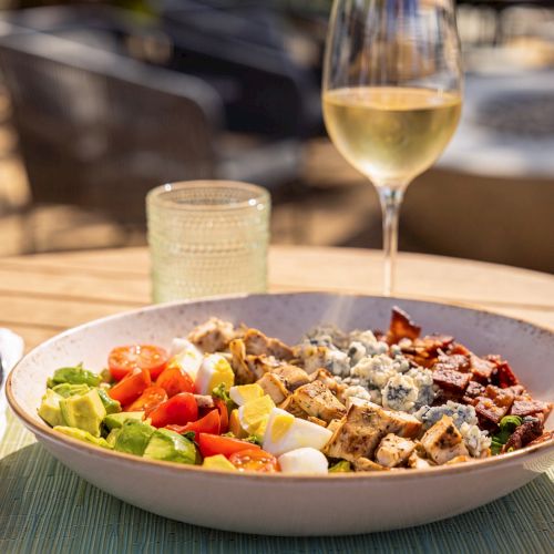 A plate of salad with greens, tomatoes, eggs and croutons sits by a glass of white wine on a patio table.