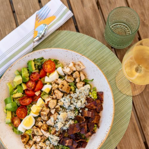 A plate of mixed salad with tomatoes, avocado, eggs, chicken, and blue cheese, served with a green drink on a wooden table.
