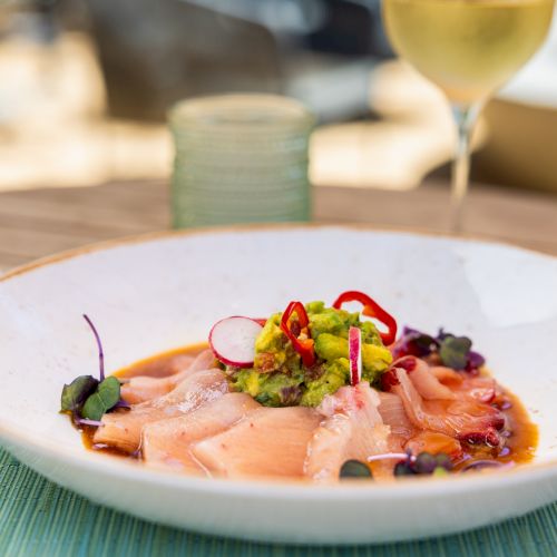 A plated salmon dish topped with avocado and colorful garnishes, with a blurred glass of drink in the background.