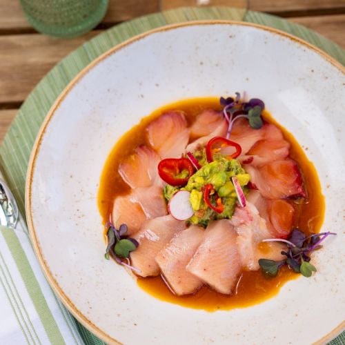 A bowl of cured salmon sashimi in orange sauce, topped with red chilies, avocado crema, radish, and microgreens.