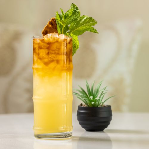 A tall glass of golden citrus drink with mint leaves and a pastry-like garnish, beside a small potted plant.