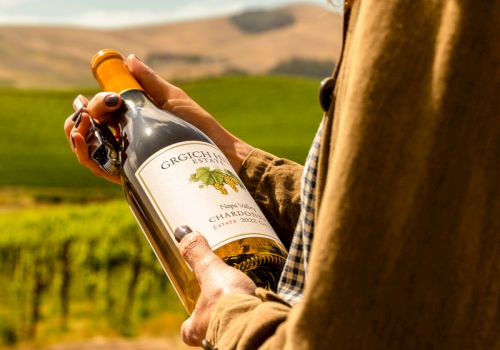 A person in a vineyard uncorks/holds a bottle of wine outdoors with hills in the background.