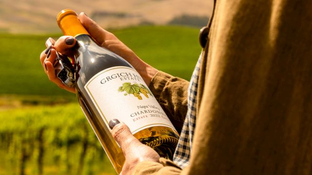 A person in a vineyard uncorks/holds a bottle of wine outdoors with hills in the background.
