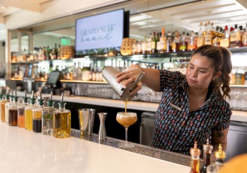 A bartender pours a caramel-colored cocktail into a glass at a bar, with bottles and a &ldquo;garnish&rdquo; sign in the background.