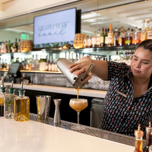 A bartender pours a caramel-colored cocktail into a glass at a bar, with bottles and a “garnish” sign in the background.