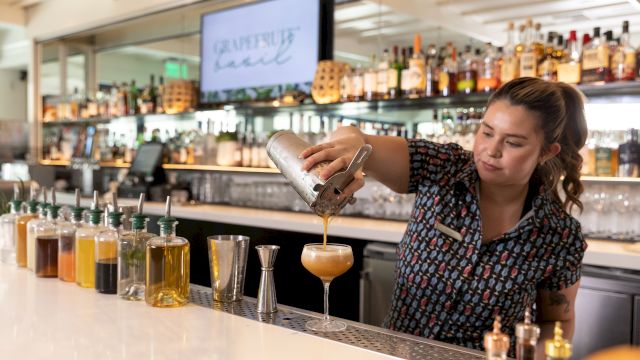 A bartender pours a caramel-colored cocktail into a glass at a bar, with bottles and a &ldquo;garnish&rdquo; sign in the background.